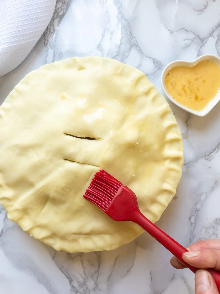 Shortcrust pastry on top of the dairy free chicken pot pie, with steam hole cut in the top, being brushed with an egg wash to turn the pastry golden