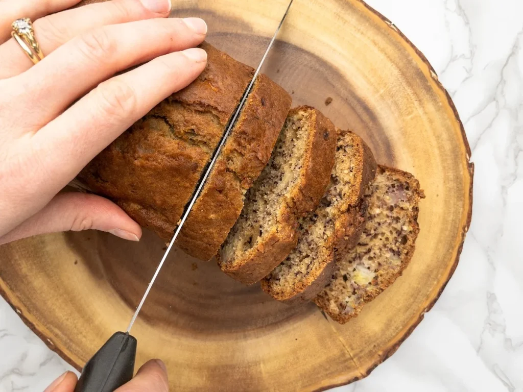 Banana bread on a wooden board being sliced with a knife.