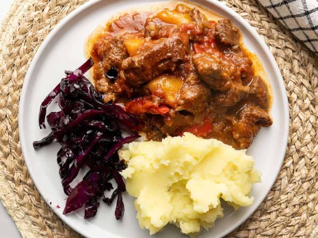 A plate of goulash served with potato and braised red cabbage.